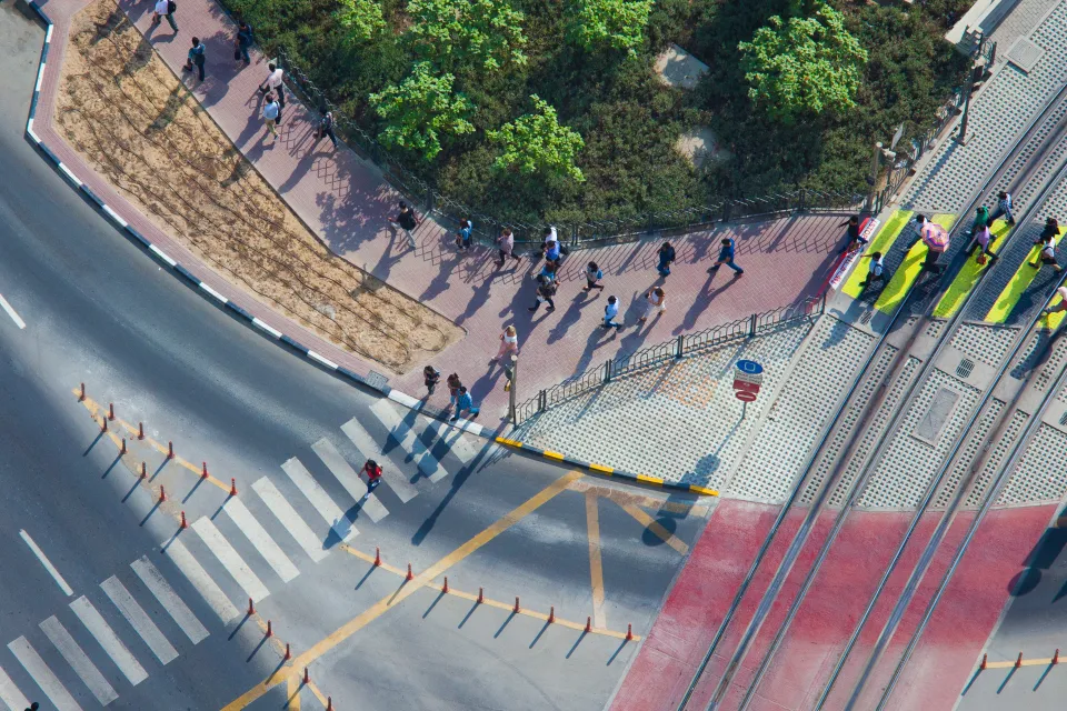 Overhead view of doxens of commuters walking on sidewalk and crossing street in full sun.
